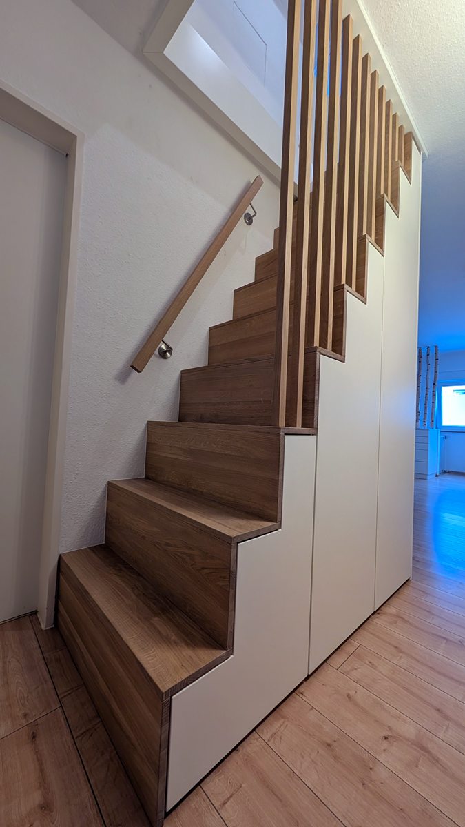 Treppe mit Stufen aus Eiche auf einem weiß lackierten Schrank mit abgestuften Türen, Eichenlamellen bis zur Decke und Handlauf an der Wand, in einem Haus in Essen