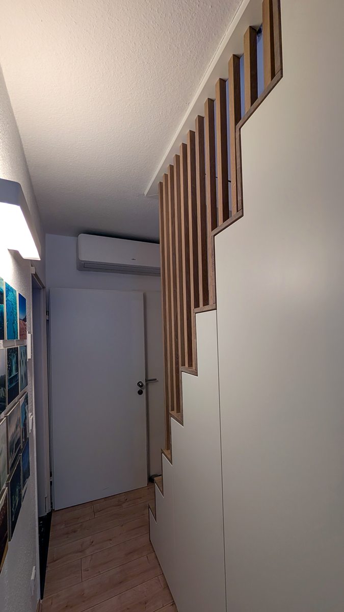 Weiß lackierter Treppenschrank mit abgestuften Türen und senkrechten Lamellen aus Eiche, vom Flur aus gesehen